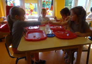 Dzieci siedzą przy stoliku, w ręce trzymają pipetki, które napełniają wodą utlenioną i przelewają do pojemników z drożdżami.